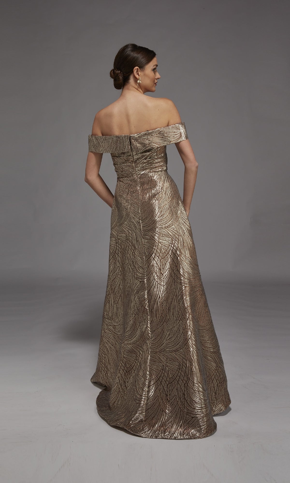 A woman stands facing away, wearing the Alyce 27844 long formal dress—an off-the-shoulder, floor-length gown with a metallic leaf-patterned jacquard design on a neutral background.